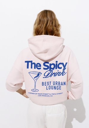 Personne aux cheveux ondulés porte un sweat à capuche rose pâle avec un texte bleu et un graphisme de verre à cocktail indiquant « The Spicy Drink, Best Urban Lounge, Los Angeles, CA. »