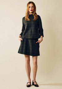 Kvinna med rött hår, klädd i en svart och grå rutig långärmad topp och matchande kjol, står med händerna i fickorna.