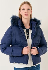 Giacca puffer blu navy con cappuccio in pelliccia sintetica, chiusura con zip e due tasche laterali. Design leggermente cropped con texture a trapunta e vestibilità aderente.