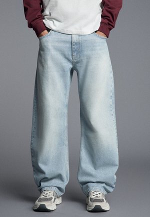 Persona che indossa jeans azzurri a gamba larga, sneakers bianche e grigie e una camicia bianca con maniche bordeaux, mani in tasca.