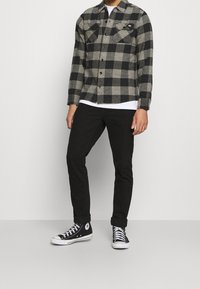 Schwarz-grau kariertes Flanellhemd mit zwei Brusttaschen, Knopfleiste, getragen über einem weißen T-Shirt, kombiniert mit schwarzen Slim-Jeans und schwarzen Turnschuhen.