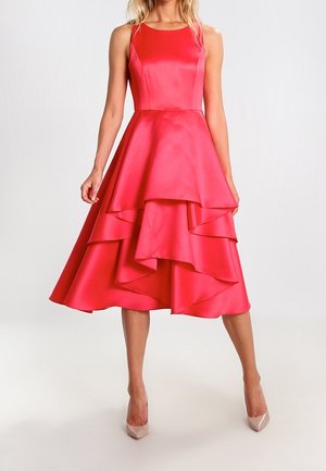 Jasnoróżowa satynowa sukienka bez rękawów z warstwową, asymetryczną spódnicą midi, dopasowanym górnym krojem, noszona z beżowymi szpilkami na jasnym tle.