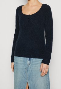 Žena nosící tmavě námořnický fuzzy pletený svetr se třemi malými knoflíky u výstřihu a světle modrou džínovou sukni s předním rozparkem.