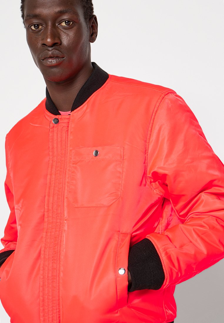Homme portant une veste bomber orange vif avec un col et des poignets noirs, les mains dans les poches, faisant face à l'avant sur un fond uni.