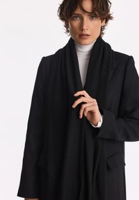 Czarny wełniany płaszcz o dopasowanym kroju, z kołnierzem klapowym, bocznymi kieszeniami oraz miękkim, zwisającym czarnym szalikiem. Gładka textura i minimalne akcenty.