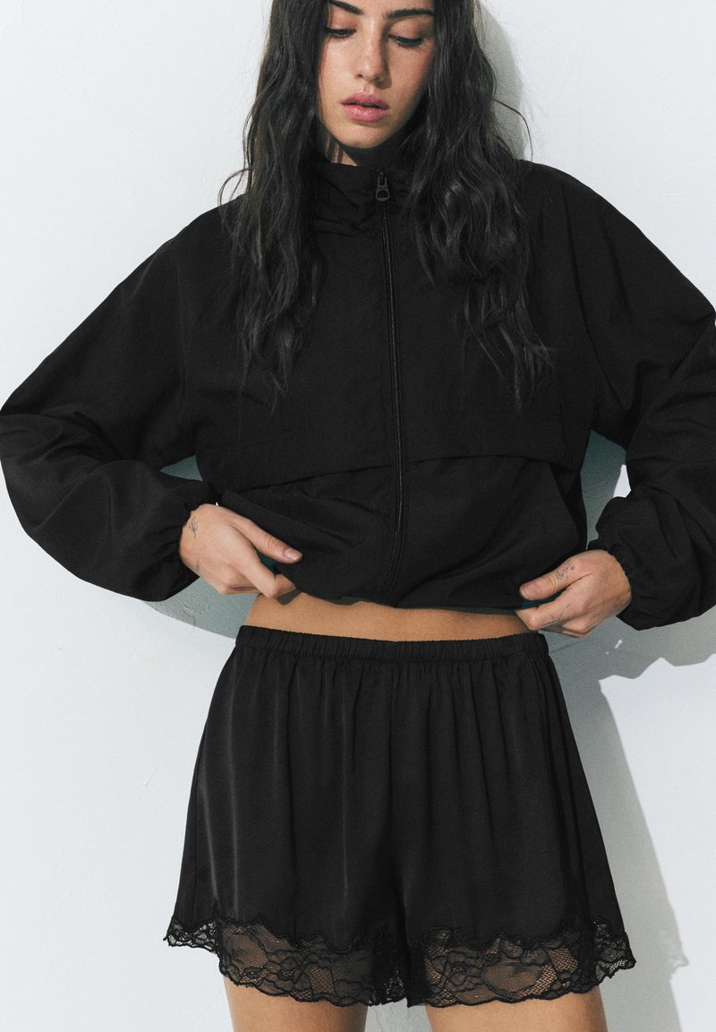 Femme aux longs cheveux foncés remontant une veste noire zippée, portant un short noir ample avec bordure en dentelle sur un fond clair uni.