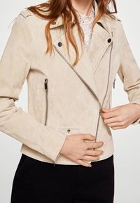 Donna che indossa una giacca in suede beige chiaro con zip e bottoni a pressione, mentre chiude la zip frontale, abbinata a pantaloni neri.
