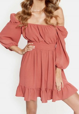 Mujer con vestido rosa polvo de hombros descubiertos, mangas abullonadas y dobladillo con volantes, posando con una mano en la cadera y la otra relajada.