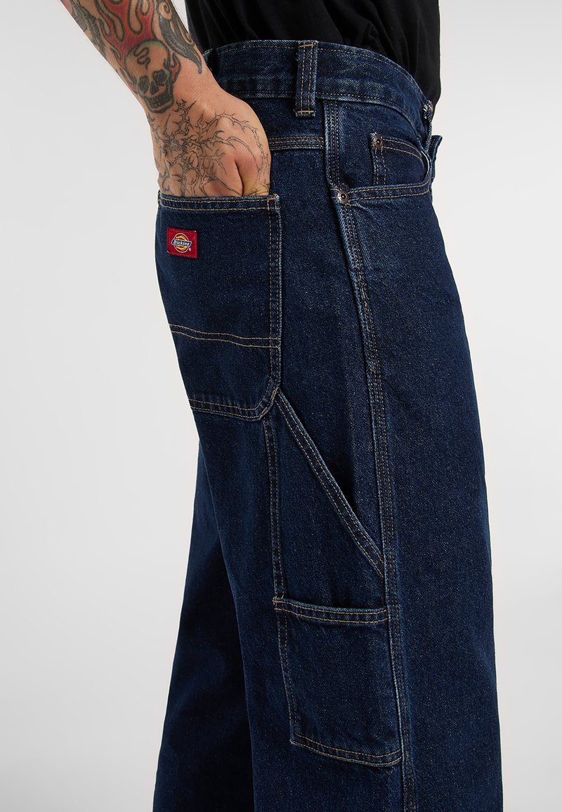 Tattooeret arm med hånd i baglommen på mørkeblå arbejdsjeans med flere lommer og en rød Dickies-logo lapel.