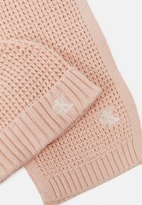 Light pink knit scarf and hat set with textured patterns and ribbed edges. Each piece features a small embroidered logo.