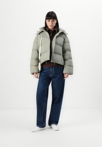 Lynette grøn pufferjakke med en cropped design, lynlås og aftagelig hætte, parret med en brun sweater og vidde bukser i blå denim. Hvide sneakers fuldender looket.