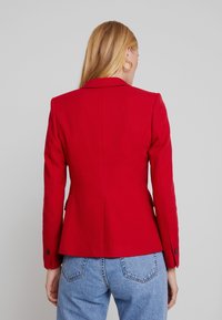 Punainen bleiseri, jossa on rakenteellinen muotoilu, teksturoitu kangas, slim fit -leikkaus, notch-lapelsit ja nappikoristeet, yhdistettynä korkeavyötäröisiin sinisiin farkkuihin.