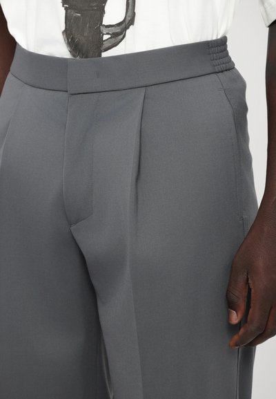 Pantaloni grigi su misura con cintura testurizzata, pieghe e tasche laterali. Il tessuto appare liscio e leggero. Mano mostrata per scala.