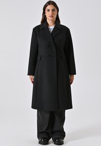 Cappotto in misto lana nera con design doppiopetto, grandi bottoni, rever a punte e una silhouette lunga e leggermente svasata.