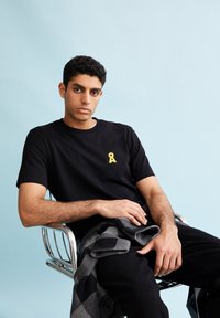 Zwarte t-shirt met korte mouwen en een geel logo, gedragen met zwarte broek. Persoon zit op een chroom stoel tegen een lichtblauwe achtergrond.