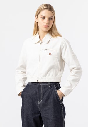 Giacca di jeans bianca cropped con zip frontale, colletto a punta e piccola tasca sul petto; indossata con pantaloni di jeans scuri con cuciture a contrasto.
