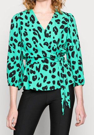 Nainen, joka pukeutunut teekinvihreään kietaisupaitaan, jossa on musta leopardi-kuvio, kolme neljäsosaa pituiset liehukkeet hihassa ja sivusolmu, yhdistettynä mustiin vartalonmyötäisiin housuihin.