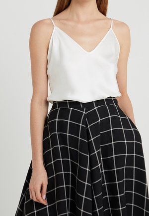 Femme portant un haut blanc sans manches à col en V associé à une jupe noire taille haute avec un motif à carreaux blancs.