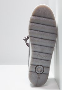 Semelle d'une basket grise Gabor avec des lignes de grip texturées et un coussin d'air circulaire, positionnée à la verticale contre un fond blanc d'angle.