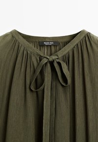Blusa verde oliva con un tessuto morbido e strutturato. Presenta una chiusura a fiocco al collo e pieghe verticali sul davanti. Etichetta del marchio visibile.