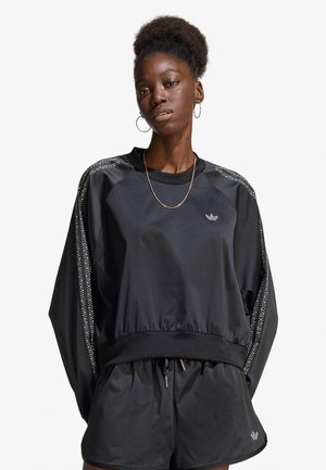 Donna con capelli naturali che indossa una felpa nera Adidas e pantaloncini con strisce bianche a pois e piccolo logo sul petto e sulla gamba.