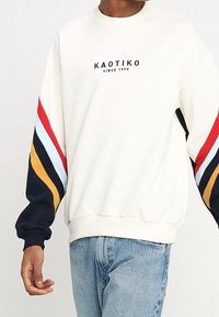 Pullover in crema con logo nero. Presenta strisce diagonali multicolori sulle maniche in rosso, blu, giallo e blu navy. Texture morbida.