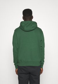 Mörkgrön hoodie med stor huva, ribbade muddar och nederkant, gjord av ett mjukt, texturerat tyg. Enfärgad design, utan synliga logotyper eller m önster.