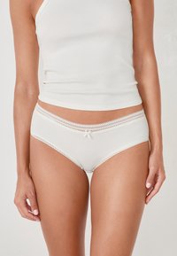 Slip in tessuto bianco a costine con bordo in pizzo in vita, caratterizzati da un piccolo fiocco al centro. Texture liscia e vestibilità aderente.