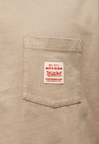 Τσέπη από μπεζ ύφασμα με ετικέτα Levi Strauss & Co. που φέρει κόκκινο κείμενο και απεικόνιση άμαξας με άλογα.