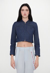Krojena modra denim jakna ima žepe v obliki srca, visok ovratnik in sprednji zadrgo, v kombinaciji s svetlomodrimi krojenimi hlačami.