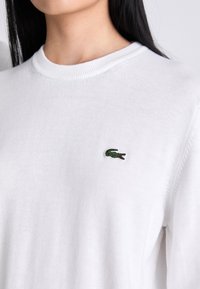 White cotton sweater featuring a round neck and ribbed edges, with a small green crocodile logo embroidered on the chest.