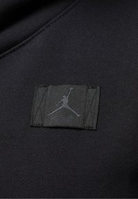 Tissu noir avec une pièce noire cousue comportant un logo Jumpman en relief et un texte en cursive subtil en dessous.