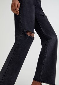 Personne portant un jean noir à jambes larges avec des déchirures usées au niveau du genou et de la cuisse supérieure, debout devant un fond uni.