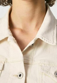 Off-white denim jacket with a classic collar, button closure, and two front pockets. Features subtle texture and stitching details.