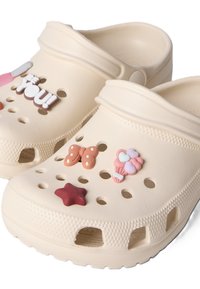 Μπεζ παντόφλες slip-on με οπές αερισμού, διακοσμημένες με γούρια που περιλαμβάνουν πεταλούδα, αστέρι, μπουκέτο μπαλονιών και κείμενο «"BE YOU!"».
