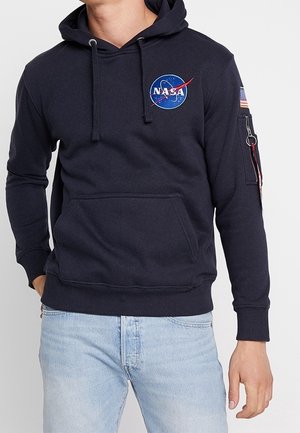 Persoon die een zwarte NASA-hoodie draagt met logo op de borst en een Amerikaanse vlagpatch op de linker mouw, gecombineerd met lichtblauwe spijkerbroek.
