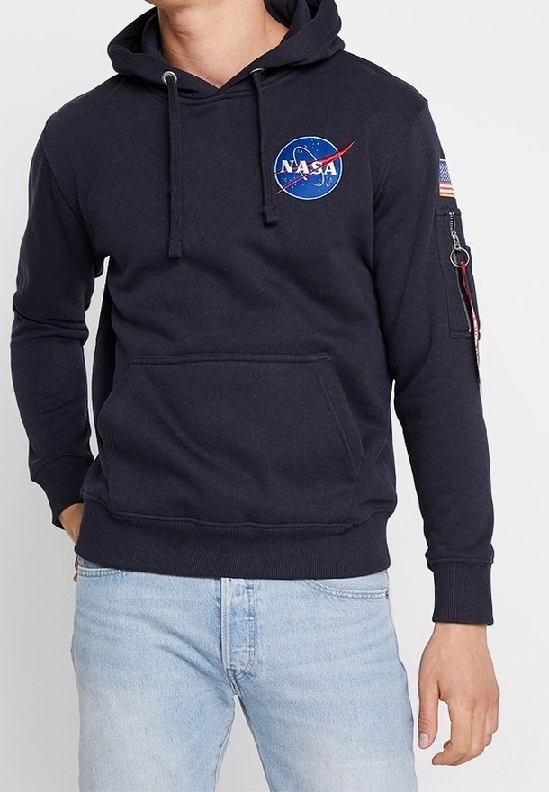 Personne portant un sweat à capuche noir NASA avec un logo sur la poitrine et un patch drapeau américain sur la manche gauche, associé à un jean bleu clair.