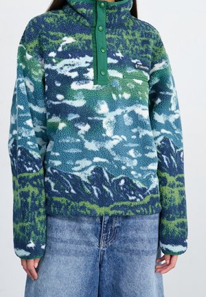 Persona que lleva un forro polar texturizado de color azul, verde y blanco con un patrón de montaña y botones de presión en el cuello, combinado con vaqueros azules.