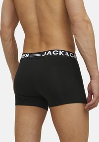 Svarta boxershorts med ett märkeselastiskt midjeband som har vit text, slät textur och figurnära design. Sedd från baksidan.