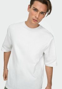 Ung mand med lys hud og brunt hår iført en almindelig hvid, oversized kortærmet skjorte mod en hvid baggrund.