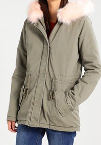 Žena oblečená v olivově zelené parce s růžovou umělou kožešinou na kapuci, předními kapsami, stahovacími pásky v pase a modrými džínami, stojící proti neutrálnímu pozadí.