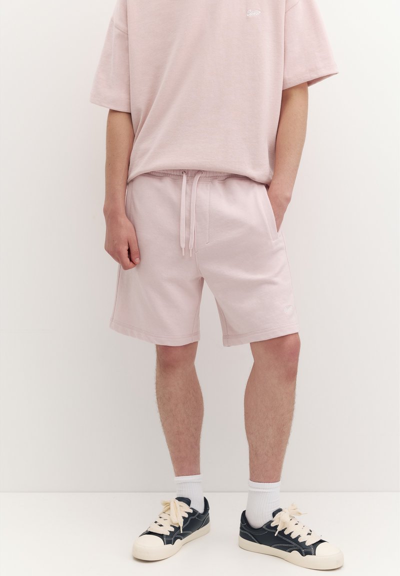 Personne portant un short rose clair et un t-shirt oversize assorti, des chaussettes blanches et des baskets basses noires avec des semelles et des lacets beiges.