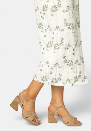 Mujer con sandalias de tacón bloque abierto de ante beige y una falda midi blanca con bordado floral gris.