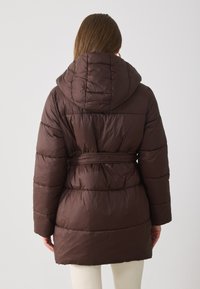 Brun puffjakke med hette, belte i midjen, quiltet tekstur og midt-i-låret lengde. Bakfra vises den glatte overflaten.