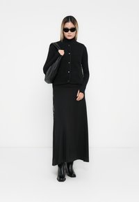 Zwarte mouwloze fleece vest over een coltrui, gecombineerd met een lange zwarte rok en kniehoge laarzen. Een slappe zwarte tas maakt de outfit compleet.