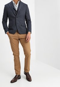 Marineblauwe gestructureerde blazer met notch-kleppen, gecombineerd met een bloemenhemd, tan chino's en bruine nette schoenen. Strak gesneden pasvorm en slank ontwerp.