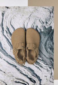 Tan suede sandals with two adjustable buckles, rounded toe, and a textured surface, displayed atop a marbled blue and white backdrop.