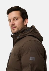 Brun polstret jakke med hætte, som har lynlås, snor og mærket patch på venstre skulder. Glat tekstur og isoleret design.