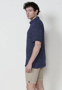 Camicia a maniche corte blu navy con tessuto testurizzato e colletto, abbinata a pantaloni corti beige chiaro con risvolto e cuciture a contrasto.