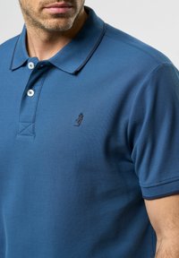 Plava polo majica od teksturiranog pamuka, sa ovratnikom, tri dugmeta i malim crnim logom na prsima.